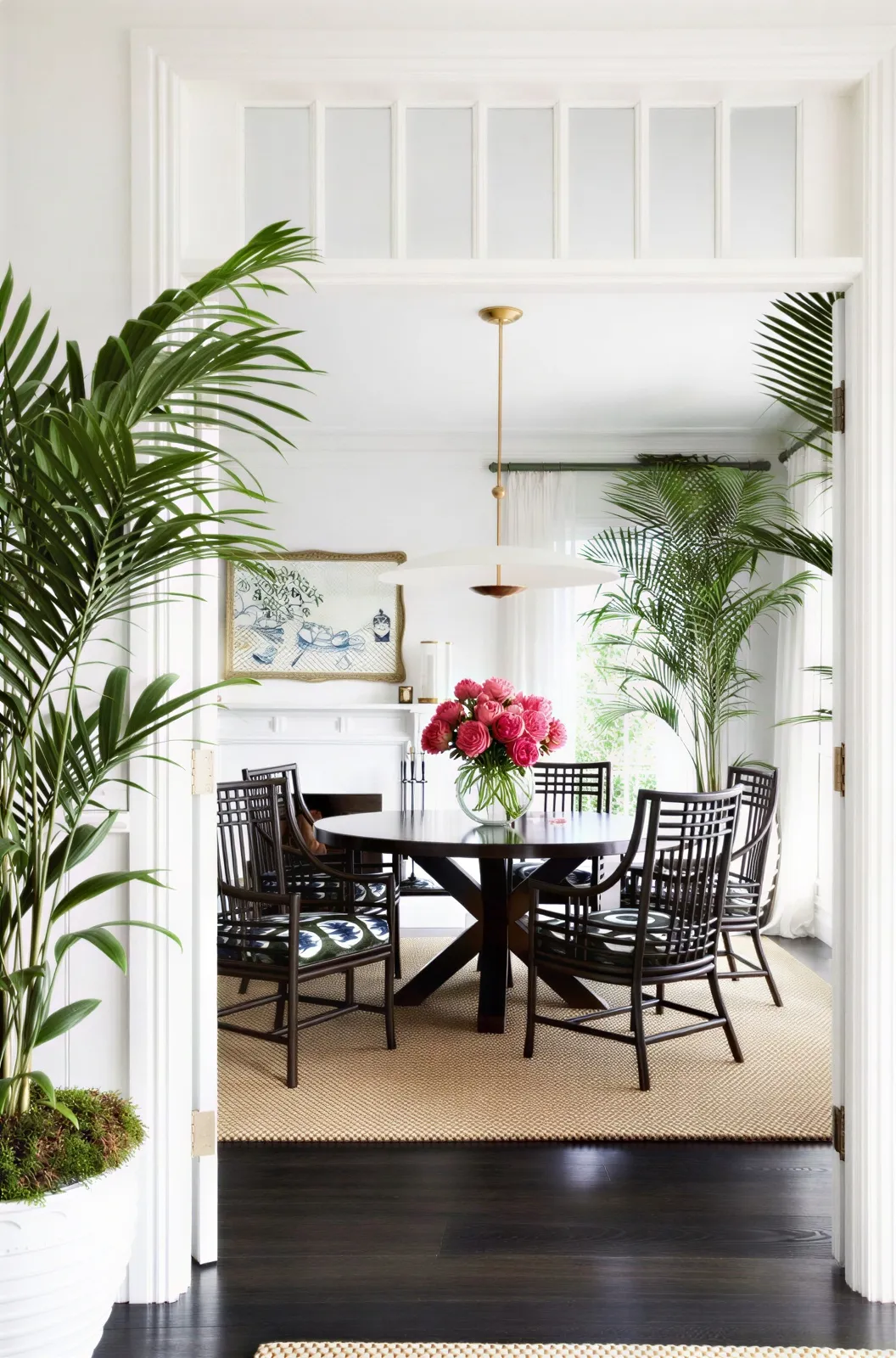 A white dining room seen through a wide doorway: near-black fretwork chairs around a dark round table, a jute rug on dark timber boards, tall palms either side of the opening and a vase of deep pink flowers on the table.