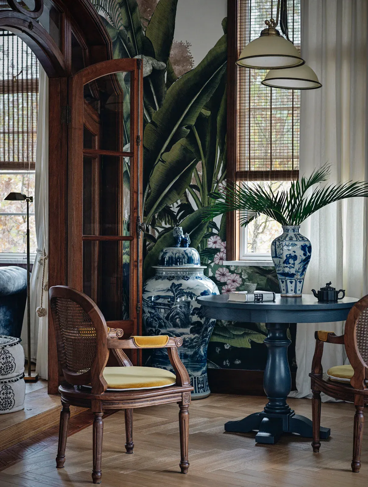 A corner of a Shanghai apartment: two cane-backed armchairs with yellow velvet cushions around a dark blue round table, a large blue-and-white Chinese ginger jar beside them, and a wall mural of banana leaves and blossom behind, with bamboo blinds at the windows.