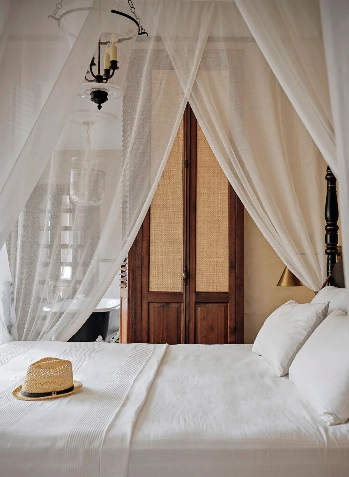 A four-poster bed with dark turned posts, draped in white mosquito netting, made up in white-on-white linen with a straw hat left on the bed. Behind it a pair of cane-panelled timber doors, and a black roll-top bath just visible through the netting to the left.