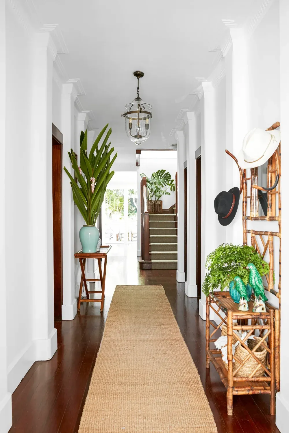 A white hallway with dark polished timber boards and a long jute runner. On the right, a tortoiseshell bamboo hallstand hung with a white panama and a black felt hat, holding a trailing maidenhair fern and two green ceramic parrots. A glass bell-jar lantern hangs overhead.