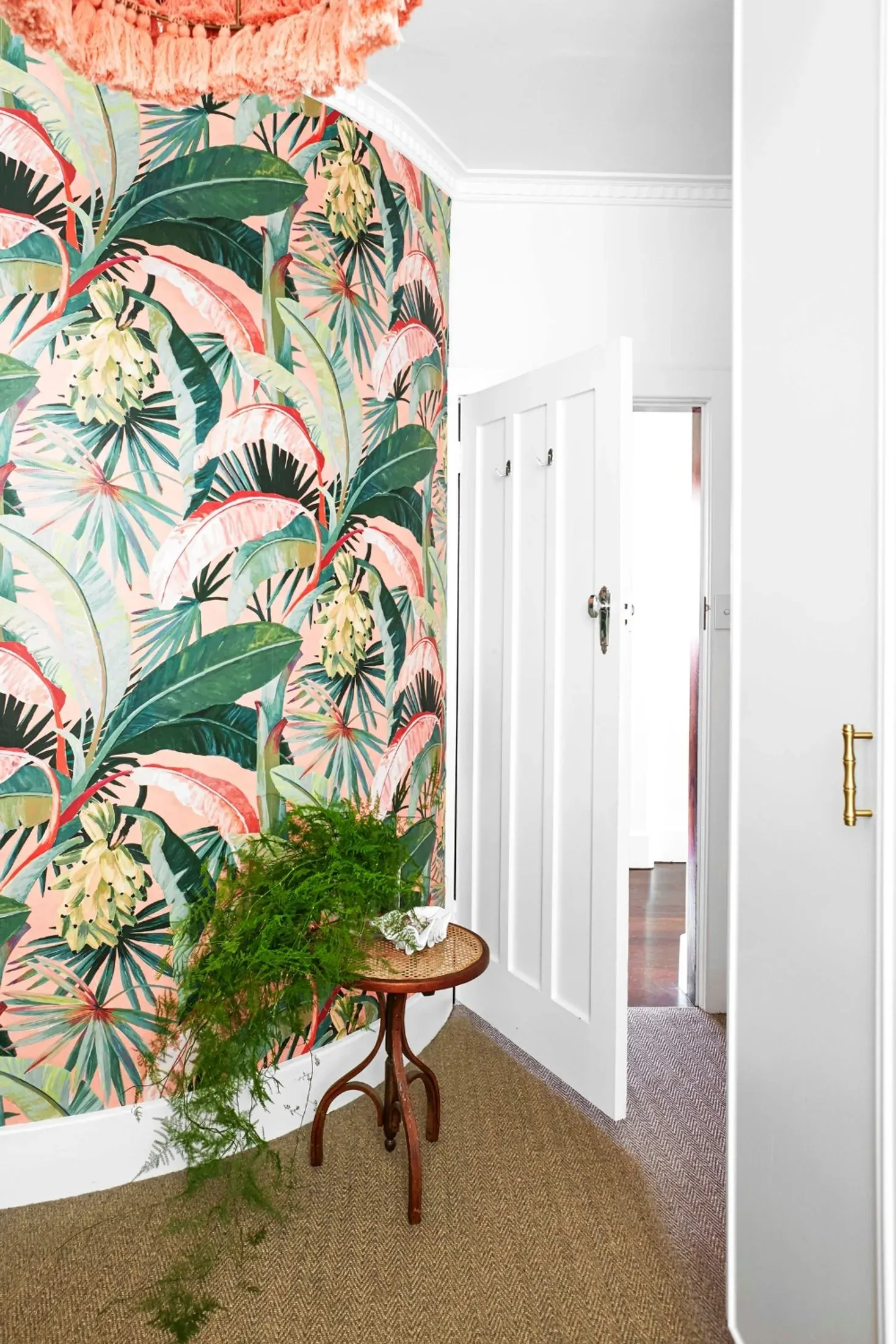 A curved wall papered in a bold tropical print of banana leaves, fan palms and pale flowers on a coral-pink ground, with a coral fringed pendant above. Against it, a small bentwood side table with a caned top holding a shell, an asparagus fern trailing to the sisal floor, and plain white joinery to the right.