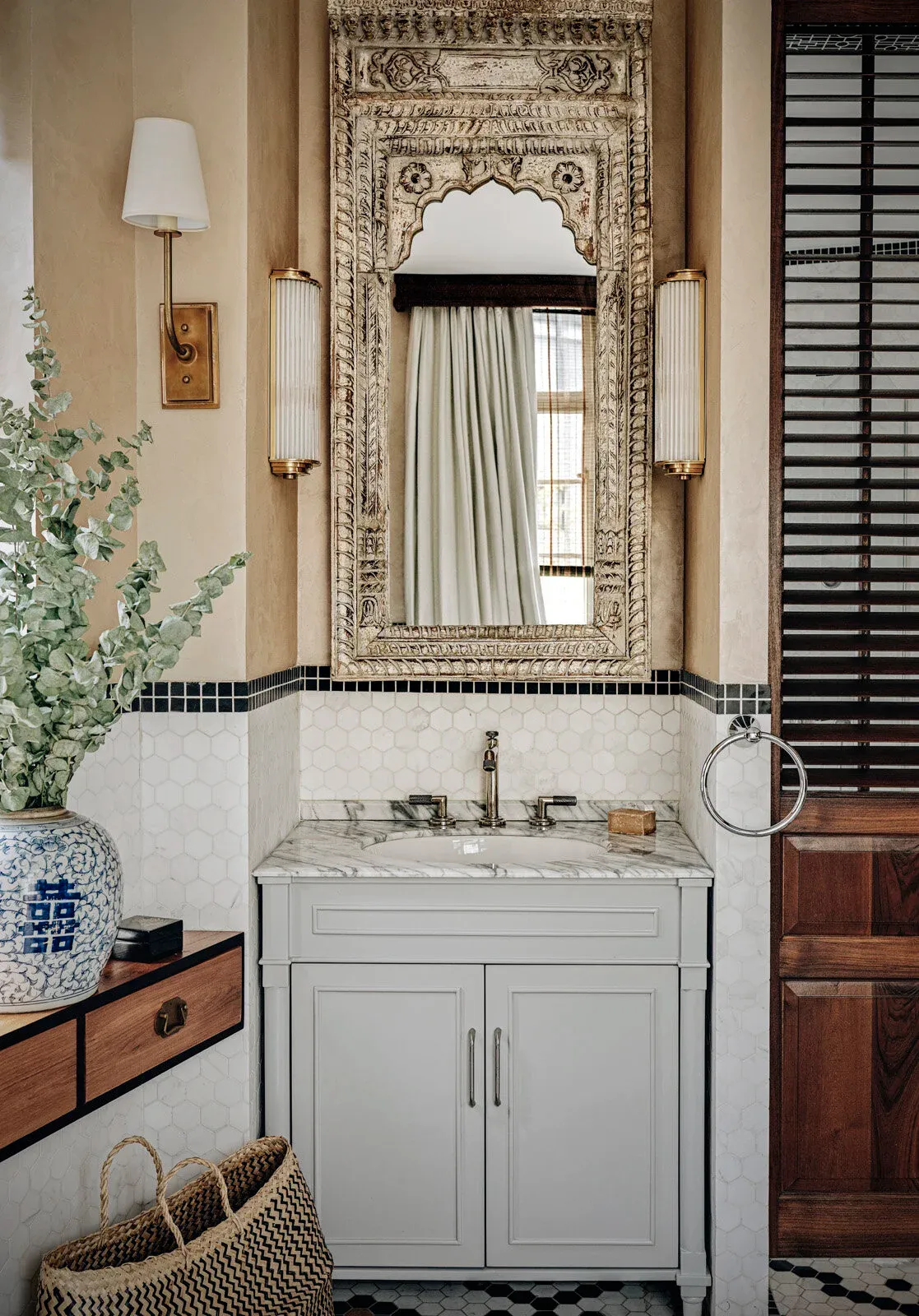 A bathroom vanity beneath a tall distressed hand-carved Indian mirror with a scalloped arch, a fluted glass and brass sconce to either side, blue-and-white porcelain on a wood console, white hexagonal tile with a dark border, and a dark timber louvred door alongside.