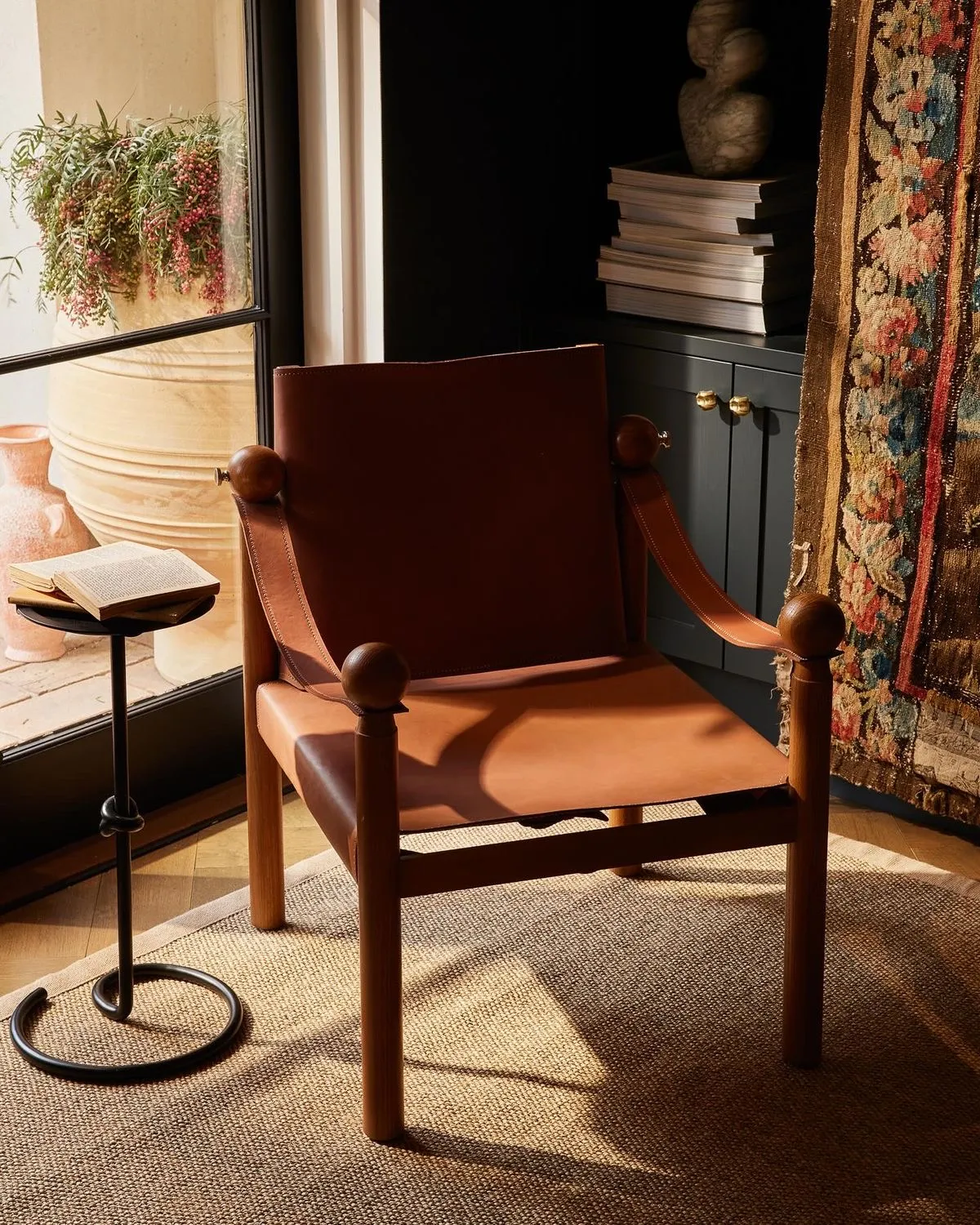 A safari chair in low afternoon sun: a tan leather sling seat and back on a turned timber frame with round ball finials, leather strap arms, standing on a sisal rug. Behind it a dark green cabinet with brass knobs, a stack of books and an antique tapestry.