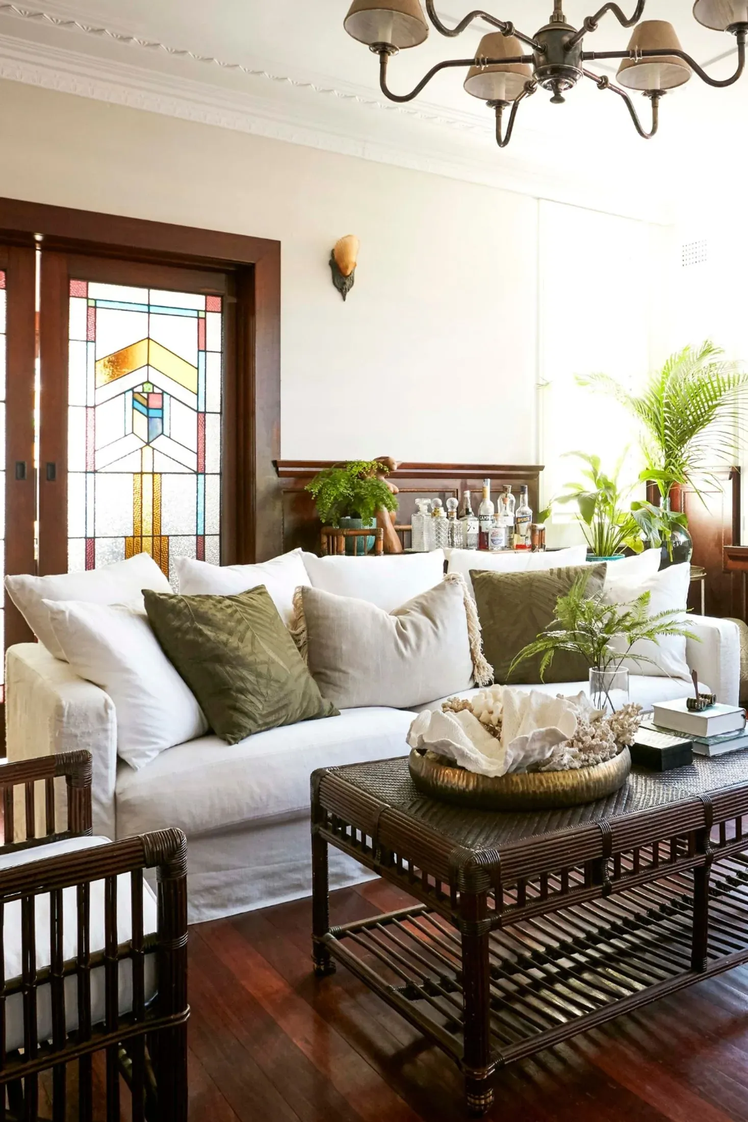 A living room with a white slipcovered sofa, a dark woven rattan coffee table holding coral in a brass tray, and a dark cane armchair, on deep red jarrah floorboards. Dark-stained timber frames an Art Deco leadlight window in amber and turquoise glass behind the sofa.