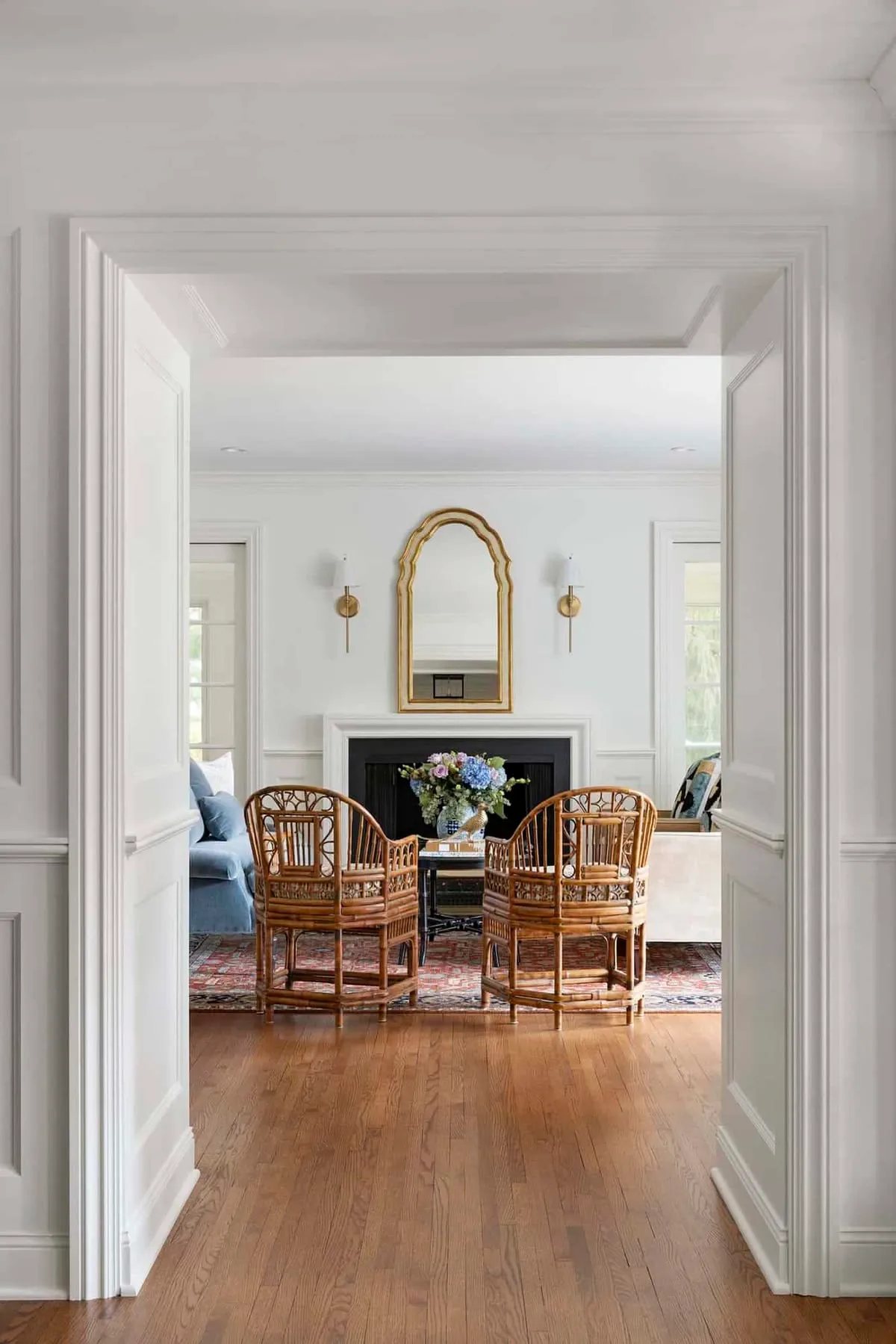 Two Brighton Pavilion cane and rattan tub chairs with rounded backs, seen through a white panelled doorway. Beyond them, a gold arched mirror above a white fireplace, a blue-and-white vase of hydrangeas, and an antique rug on oak floors.