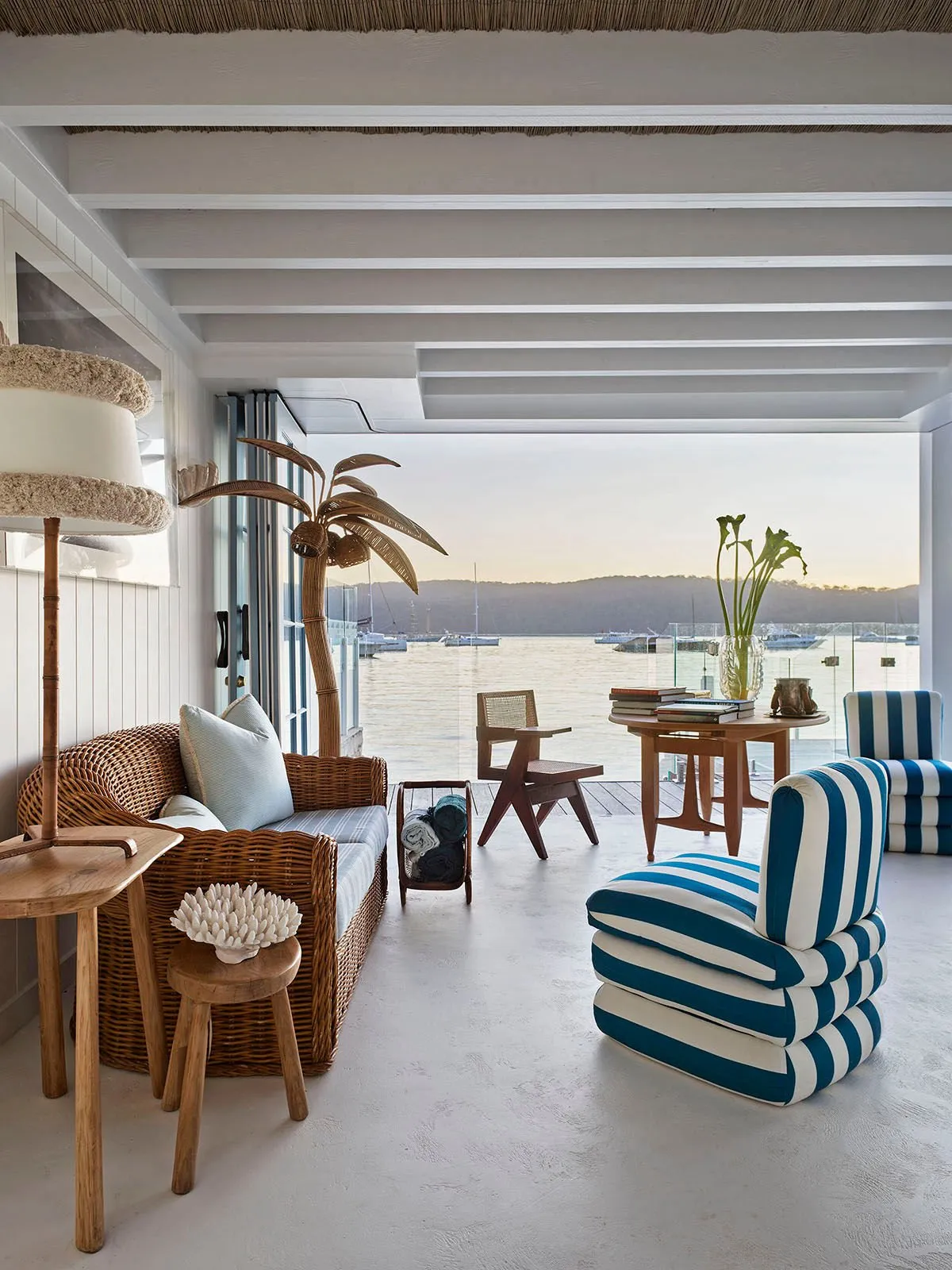 A boathouse living room with a wicker sofa, blue-and-white striped chairs and a brass palm-tree lamp, opening to a water view over a pale poured floor