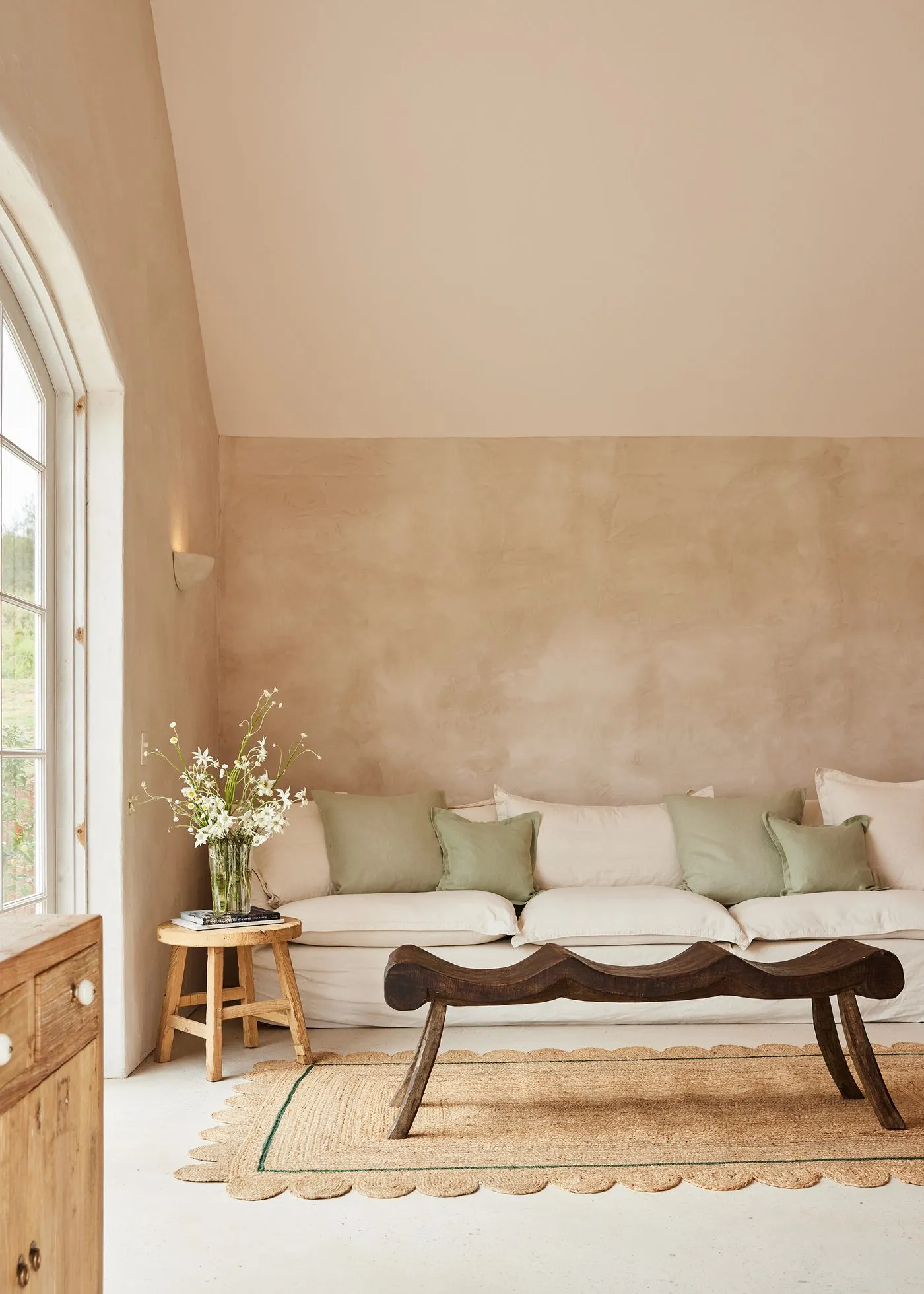 A warm plaster living room with a cream linen sofa, sage cushions and a jute rug beside an arched window