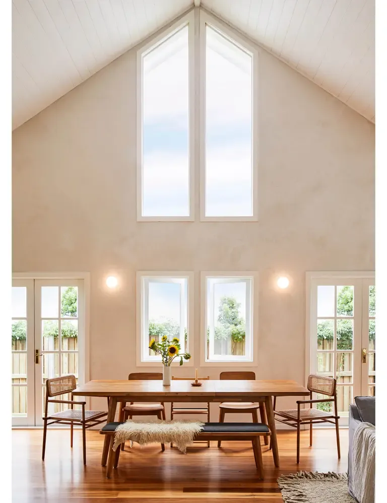 A dining room where pale plaster runs from the walls up into a high vaulted ceiling, the whole gable softly luminous before a single lamp is lit.