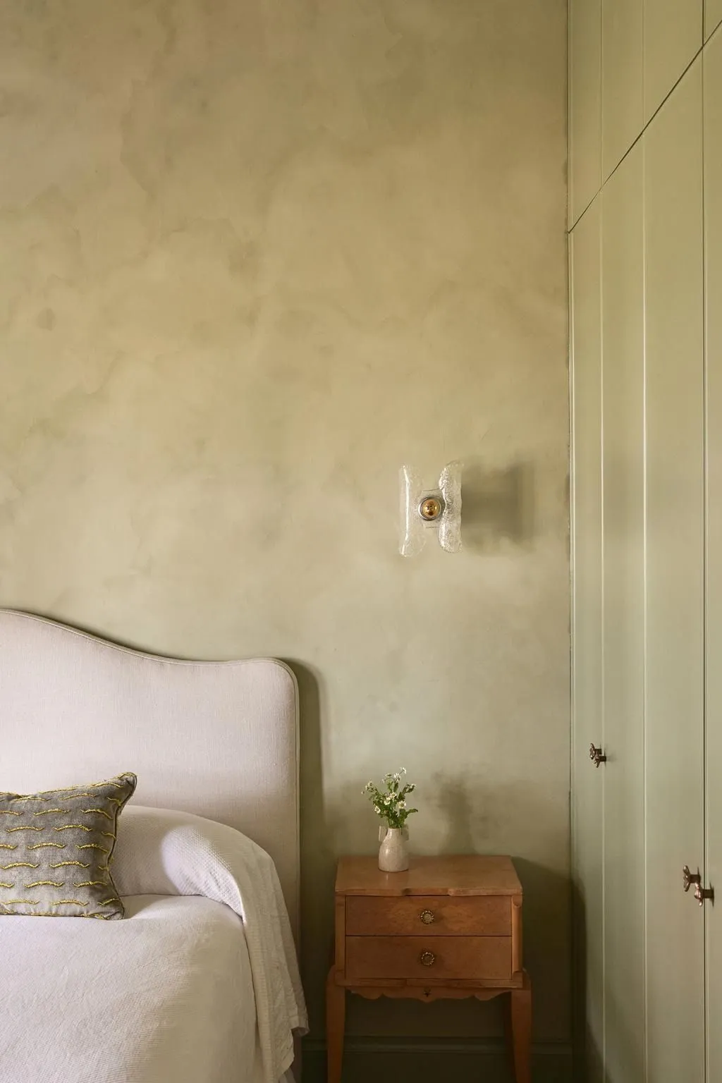 A plain linen bed against a wall limewashed in a soft, chalky green so pale it reads almost as light.